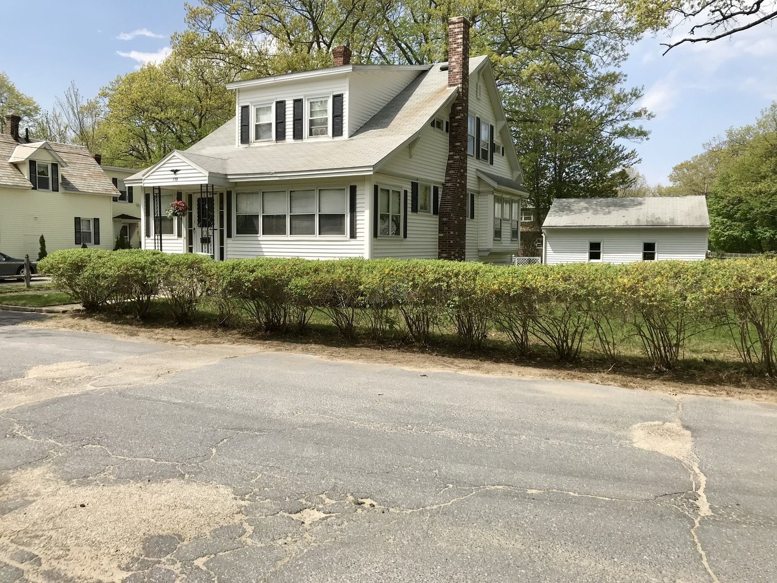White New England house behind a long roadside hedge row awaiting a trim